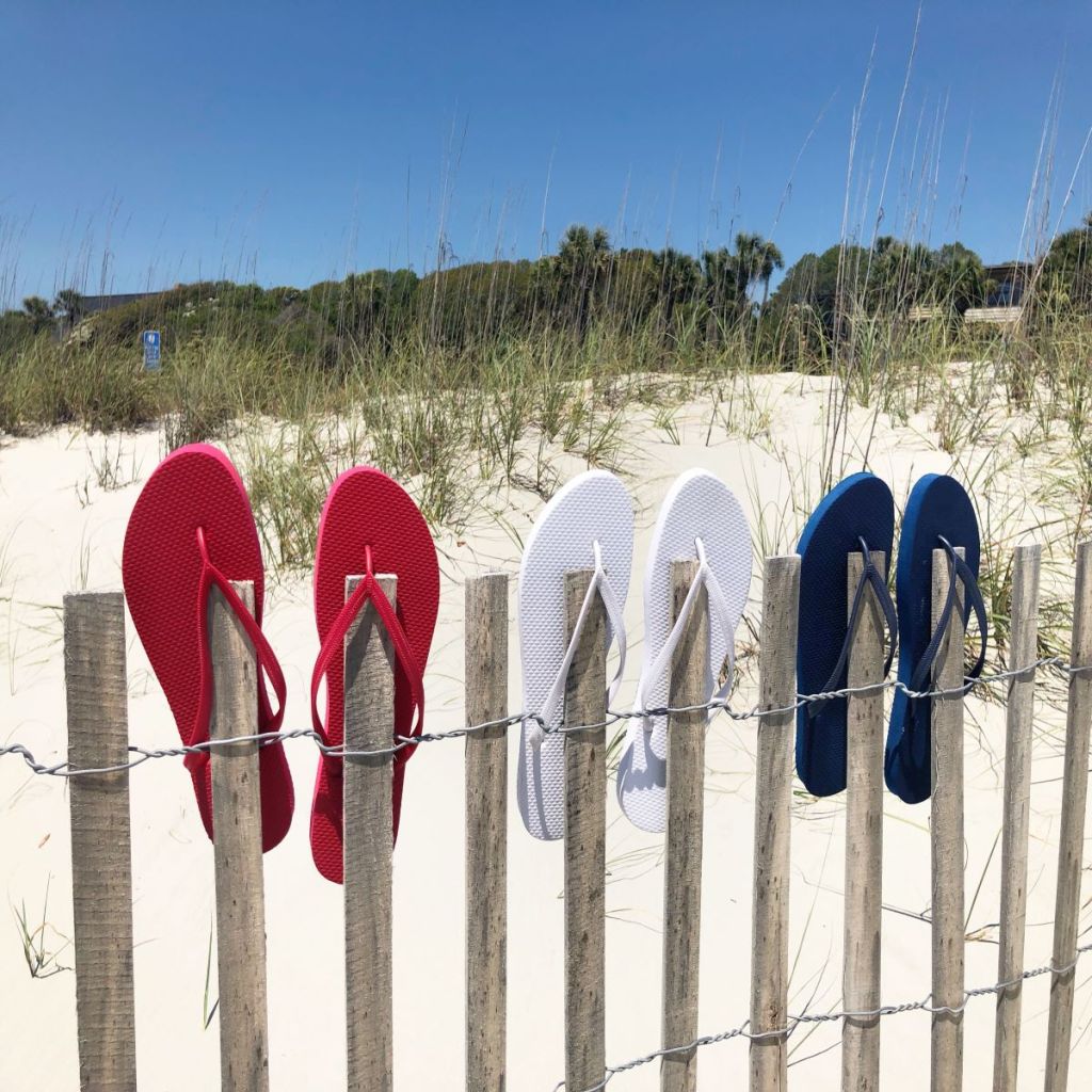 Red, white and blue flip flops on a dune fence. Sand and green trees in the background.