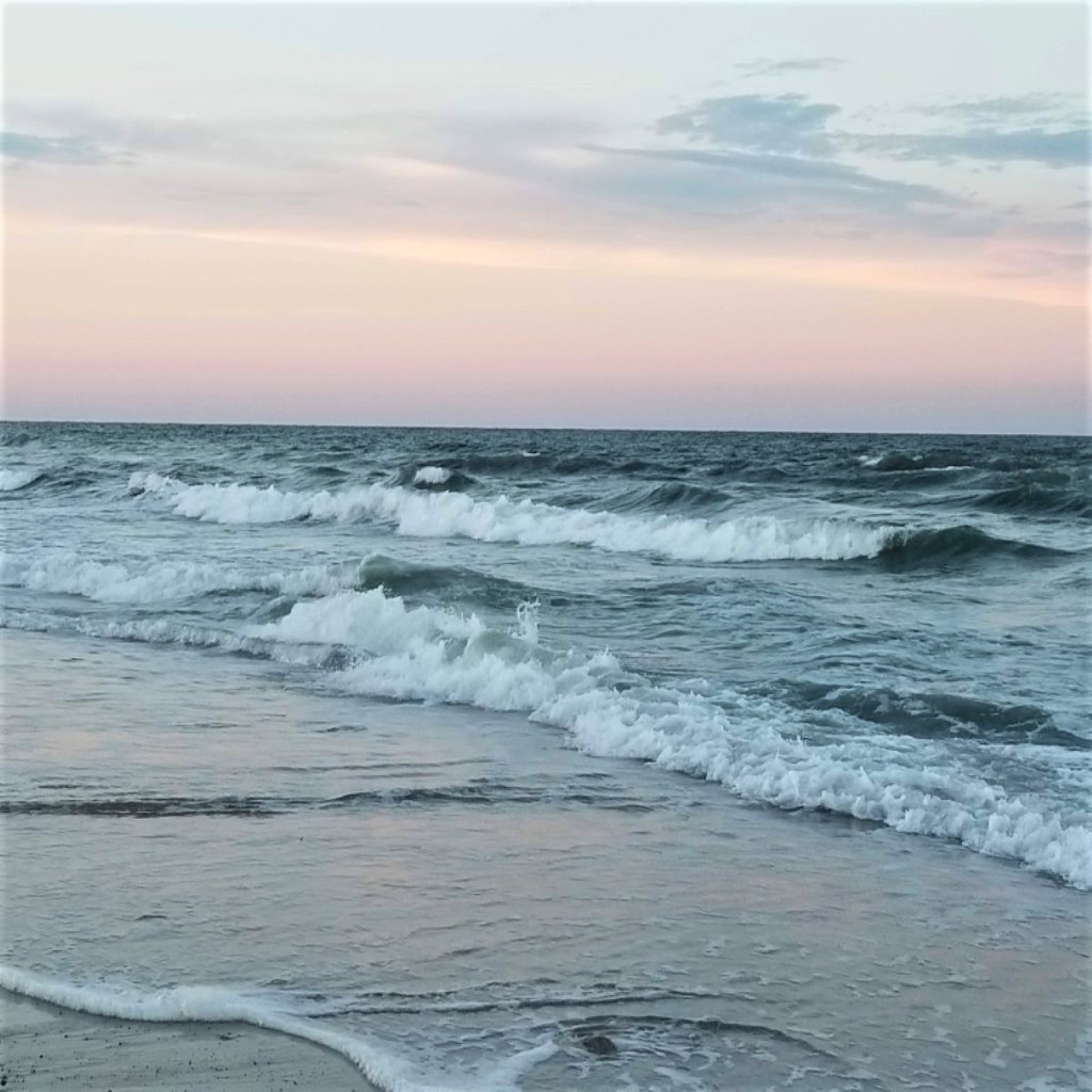 Dark blue ocean waves with white foam pink and light blue sky.
